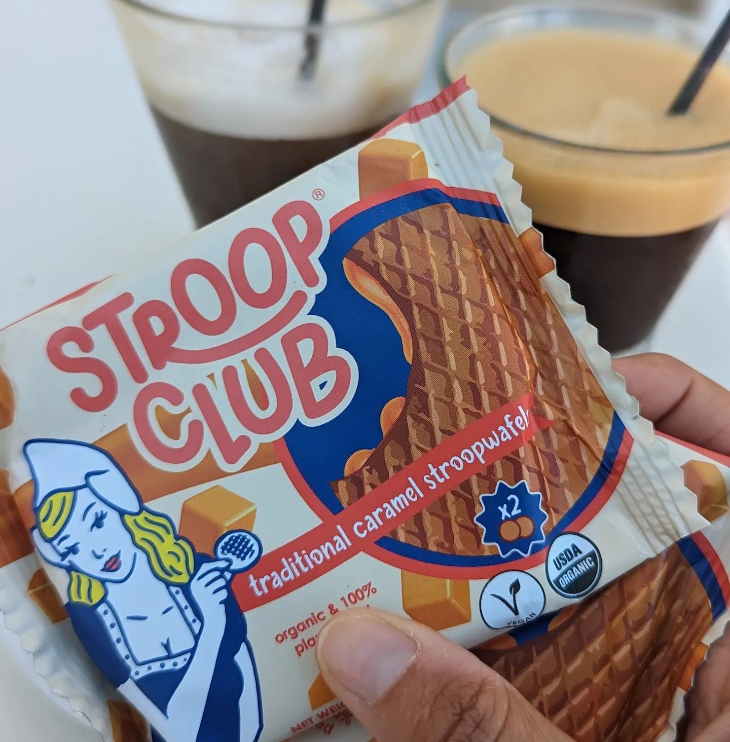 Photo of a stroopwafel 2-pack being handed, in front of coffee drinks.